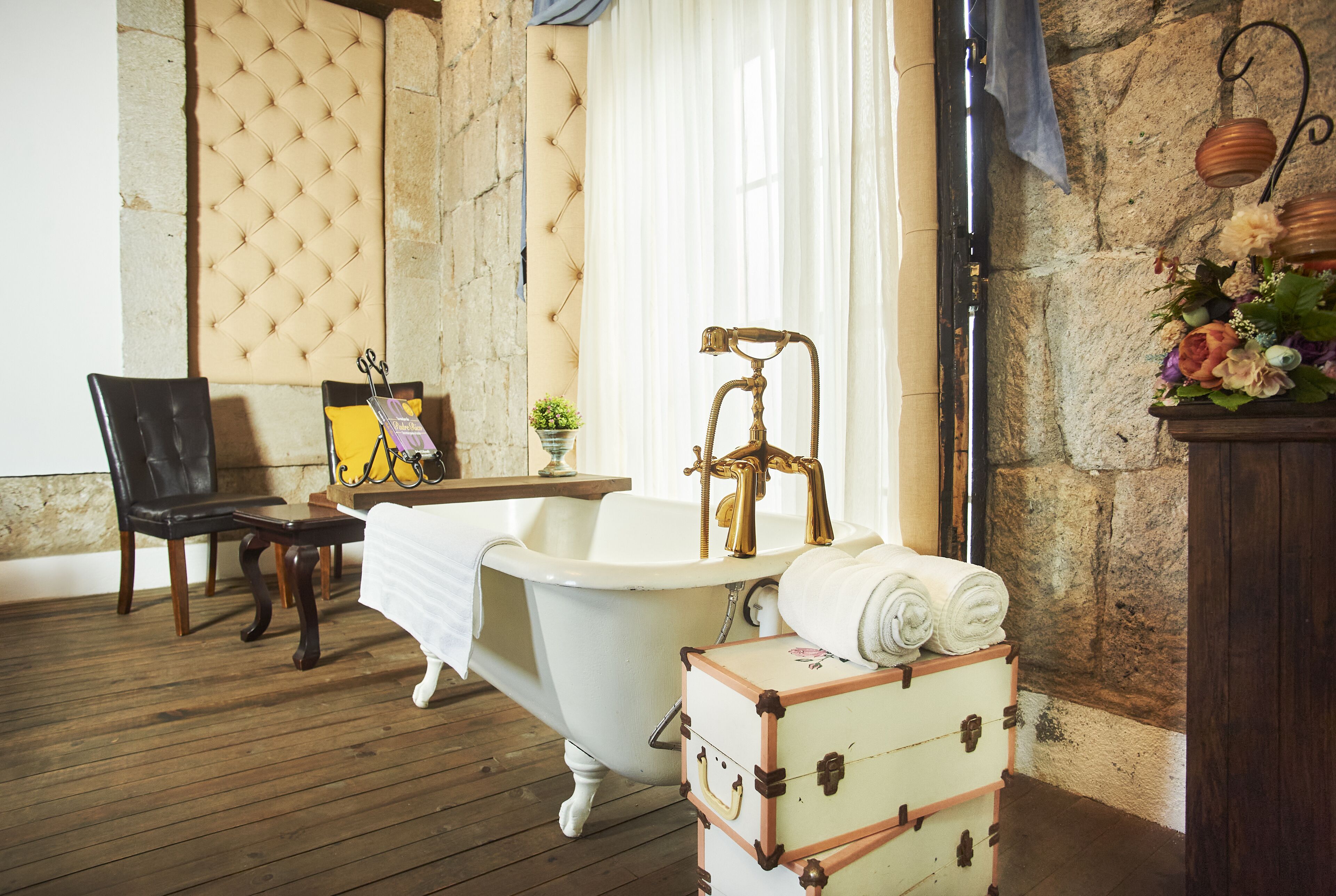 Casa de banho antiga com uma banheira de patas de leão, um banco de madeira, uma mesinha com um livro e uma mala.