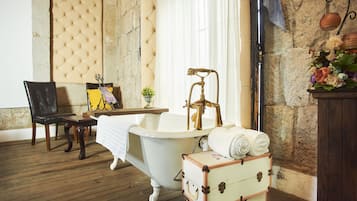 A vintage bathroom with a clawfoot bathtub, a wooden bench, a small table with a book, and a suitcase.