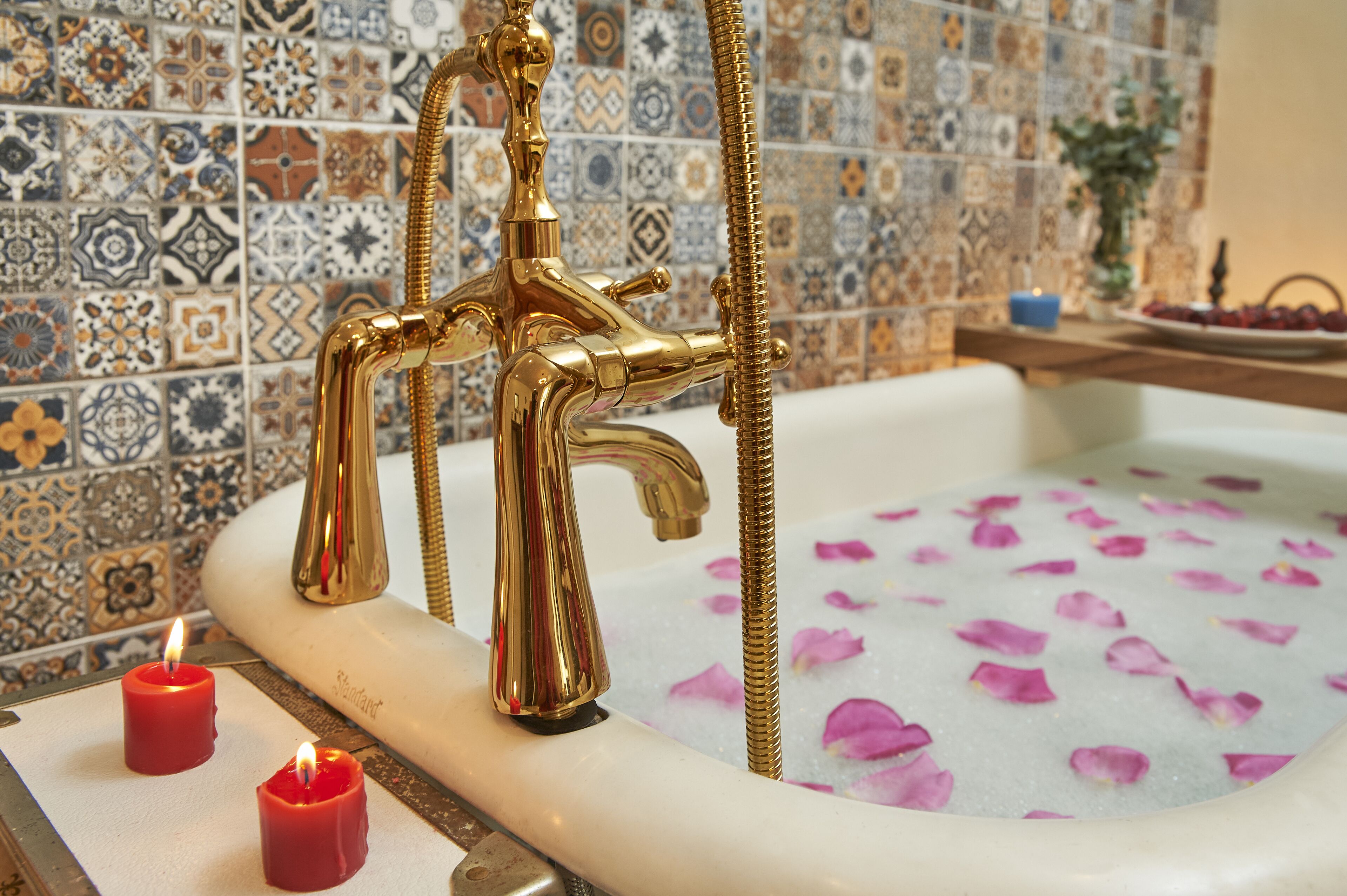 A bathtub with gold fixtures and rose petals in the water, surrounded by a tiled wall with a decorative pattern.