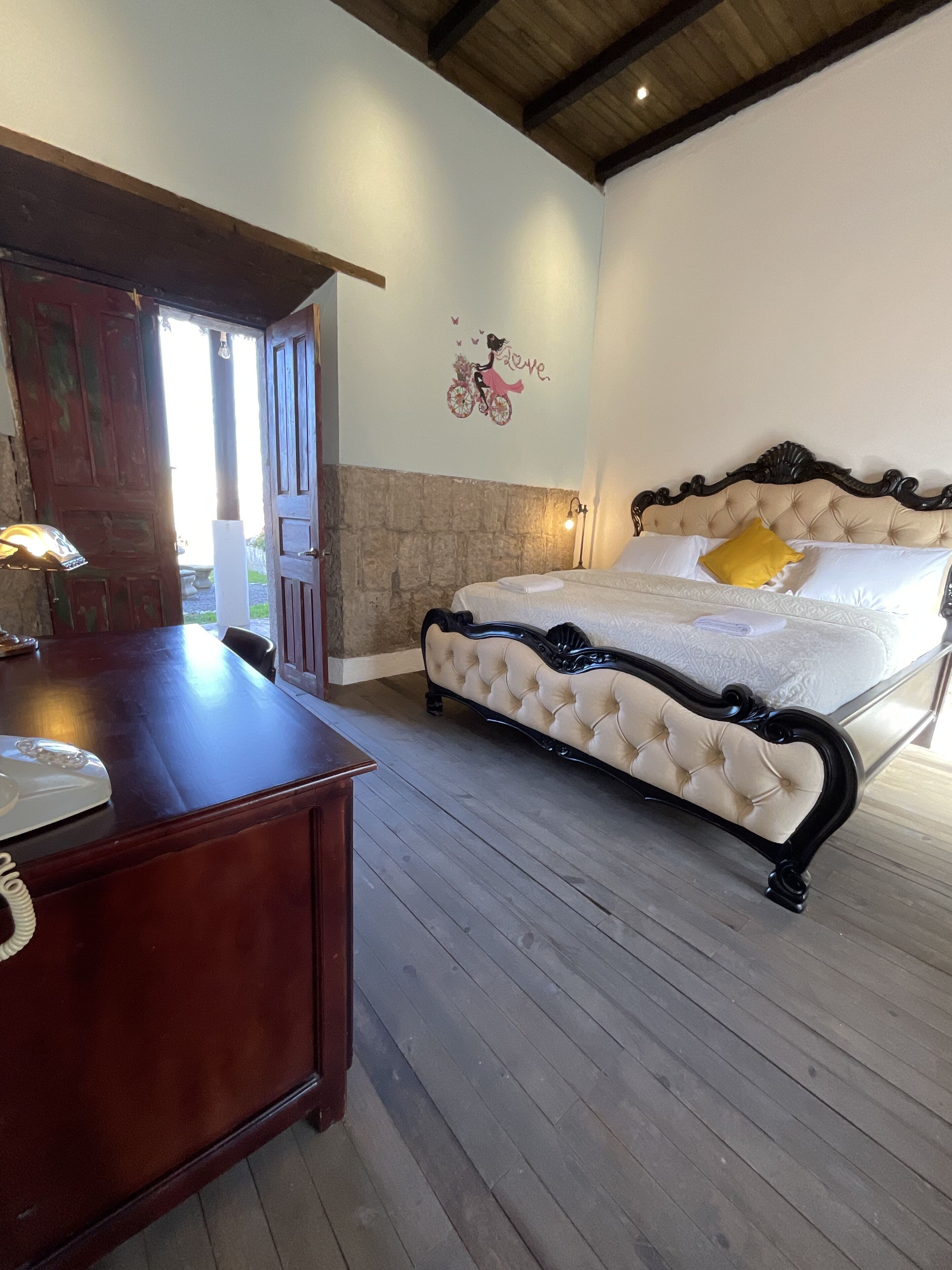 A bedroom with a wooden floor, a bed with a tufted headboard, a desk with a telephone, and a wall with a decorative sticker.