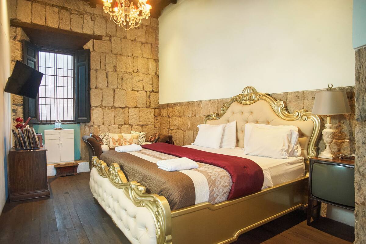 A bedroom with a large bed, stone walls, a chandelier, a television, and a small cabinet.