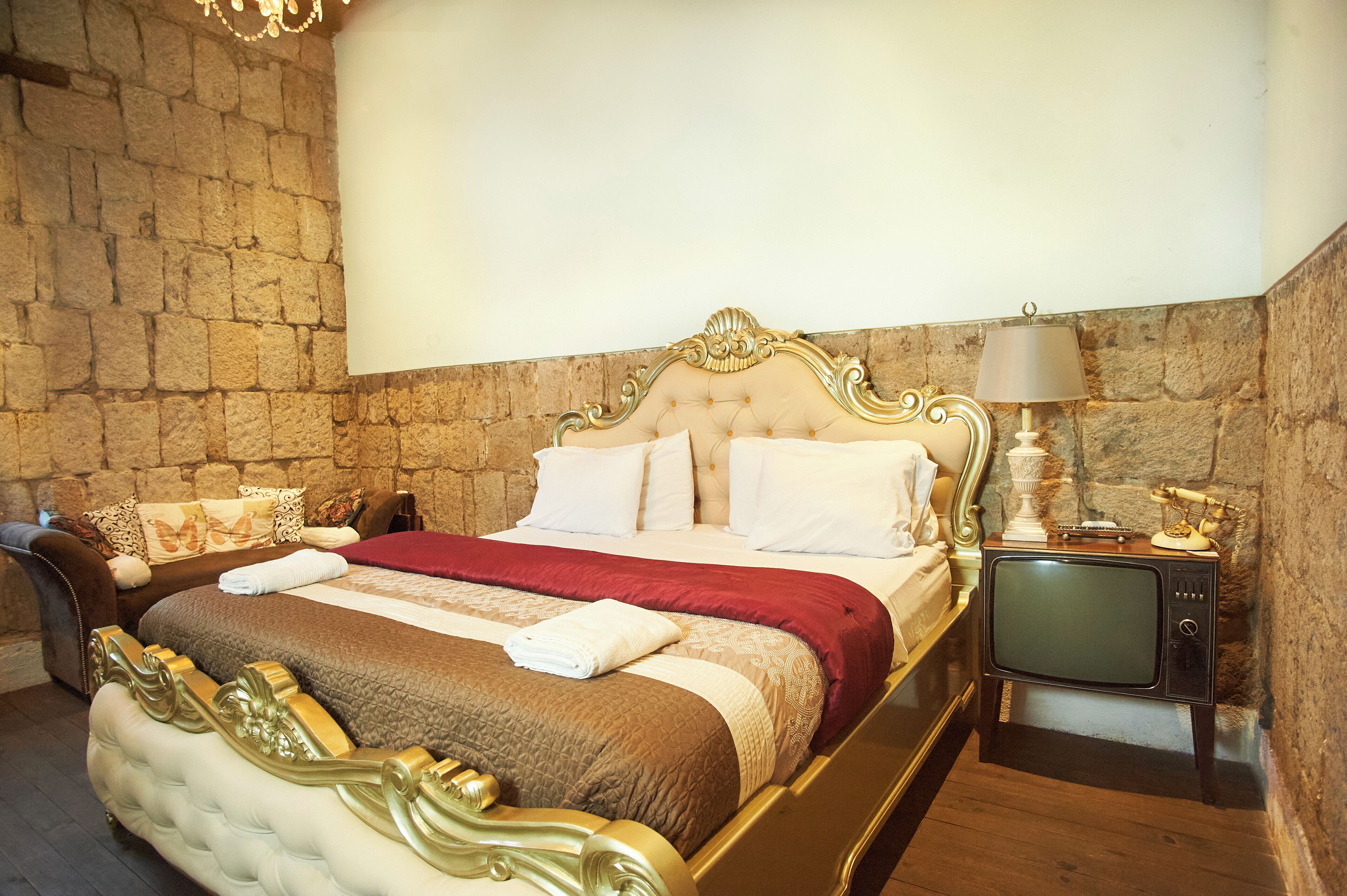 A bedroom with a stone wall, a bed with a red and brown bedspread, a television, and a lamp.