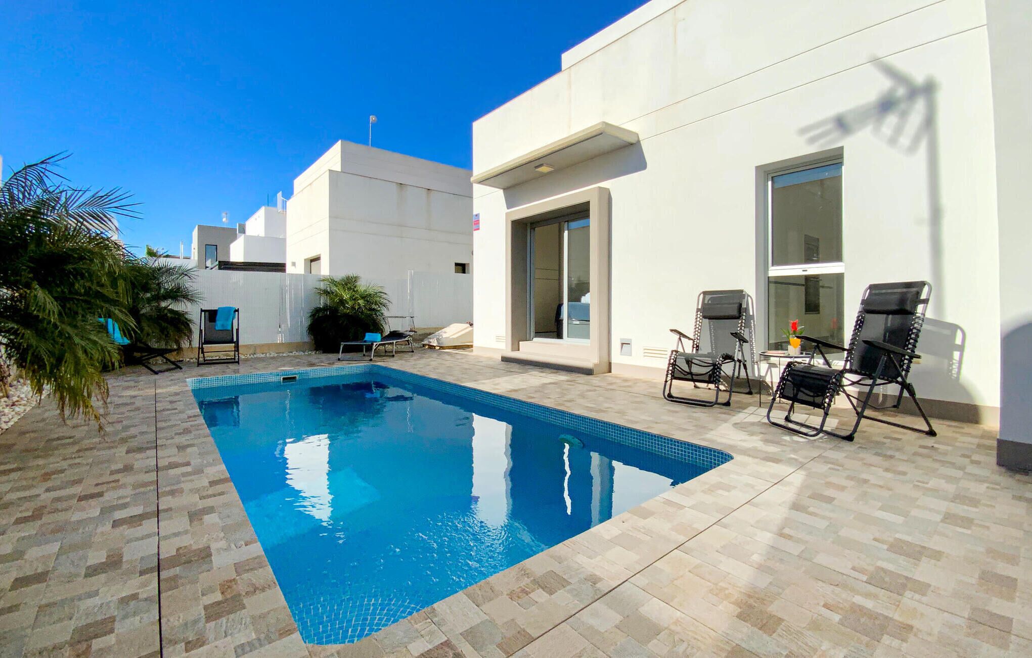 A swimming pool with tiled flooring, a patio area with outdoor furniture, and a modern building with large windows and a door.
