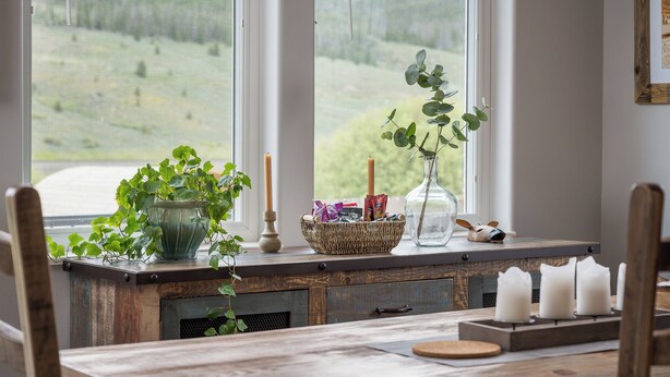 A rustic wooden table with a basket of snacks, a green plant, and candles by a window with a view of a green landscape. - Modern Home w/ Great Private Views, Hot Tub & Sauna (Tabernash)