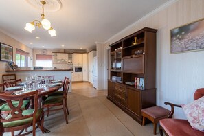 A dining area with a wooden table set for four, surrounded by chairs with green cushions. A wooden bookcase with glass doors is to the right.