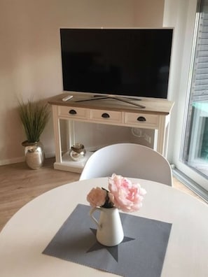 Un salon moderne avec un téléviseur à écran plat posé sur un meuble en bois, une chaise blanche, une petite table sur laquelle se trouve un vase de fleurs et une plante en pot.