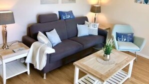 A modern living room with a dark grey sofa, a light blue armchair, a wooden coffee table, and a side table with a lamp. There are two wall-mounted picture frames and a small potted plant on the coffee table. - Möwennest N°3 - Ground floor apartment with fantastic sun terrace and garden. (Grömitz)