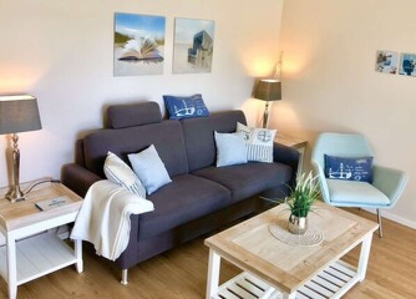 A modern living room with a dark grey sofa, a light blue armchair, a wooden coffee table, and a side table with a lamp. There are two wall-mounted picture frames and a small potted plant on the coffee table. - Möwennest No. 3 - Ground-floor apartment with a fantastic sunny terrace and garden. (Grömitz)