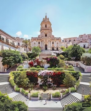 A historic building with a central staircase leading to a bell tower, surrounded by lush gardens and a symmetrical layout.