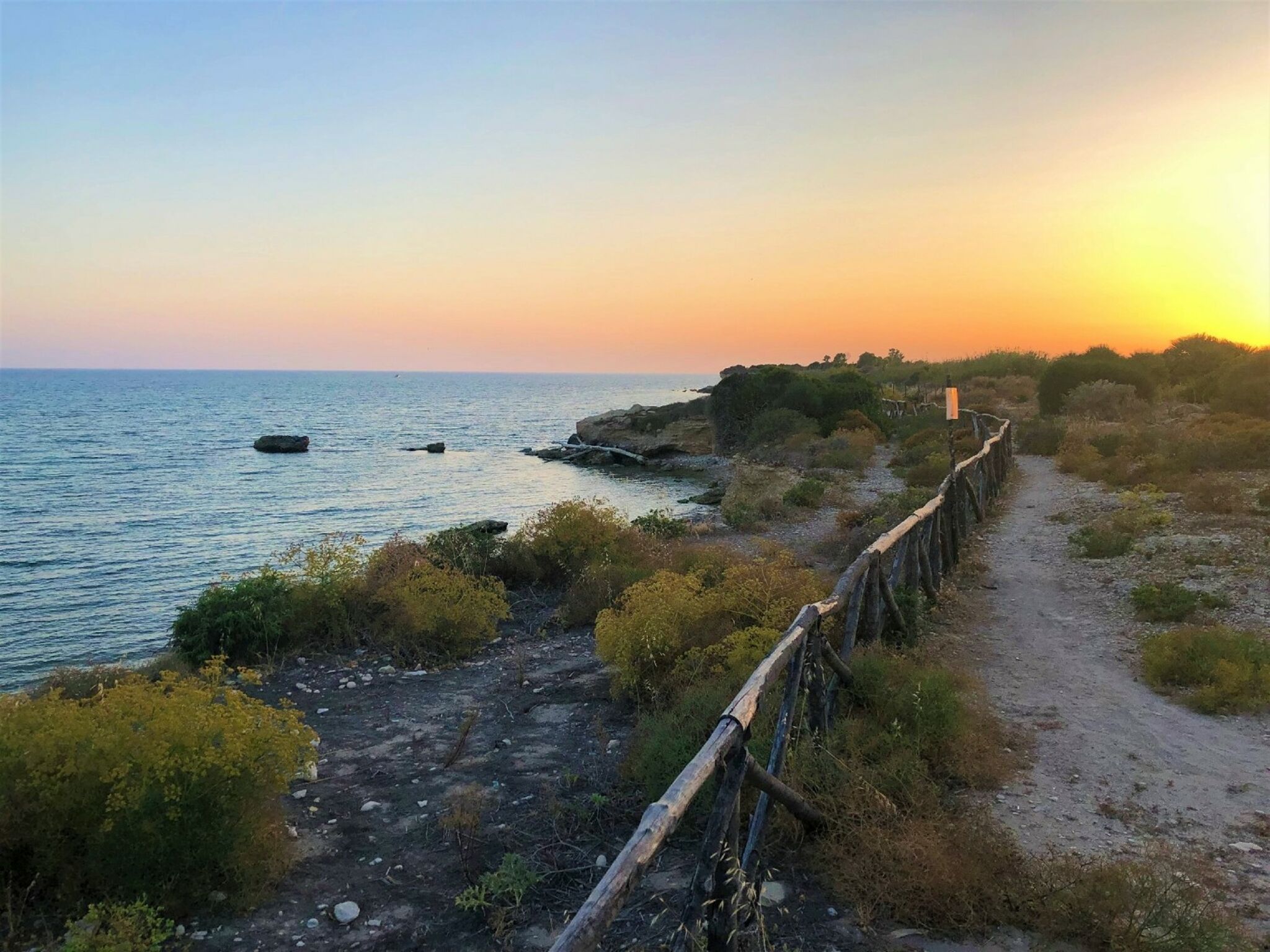A coastal path with a wooden fence, rocky terrain, and vegetation.