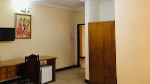 Una habitación con ventilador de techo, un escritorio con cajones, una silla, un refrigerador pequeño y un armario.
