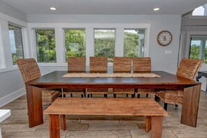 A dining area with a large wooden table and wicker chairs, large windows with a view of greenery, and a wall clock. - Large home with golf course view, open concept in living room with balcony (Bald Head Island)