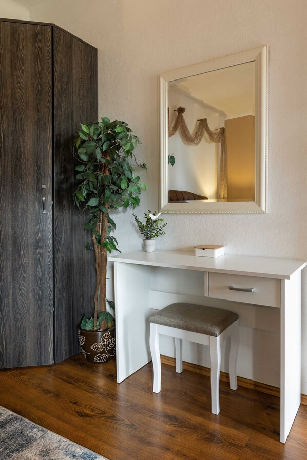 A room with a white desk, a mirror, a potted plant, and a dark wooden wardrobe.