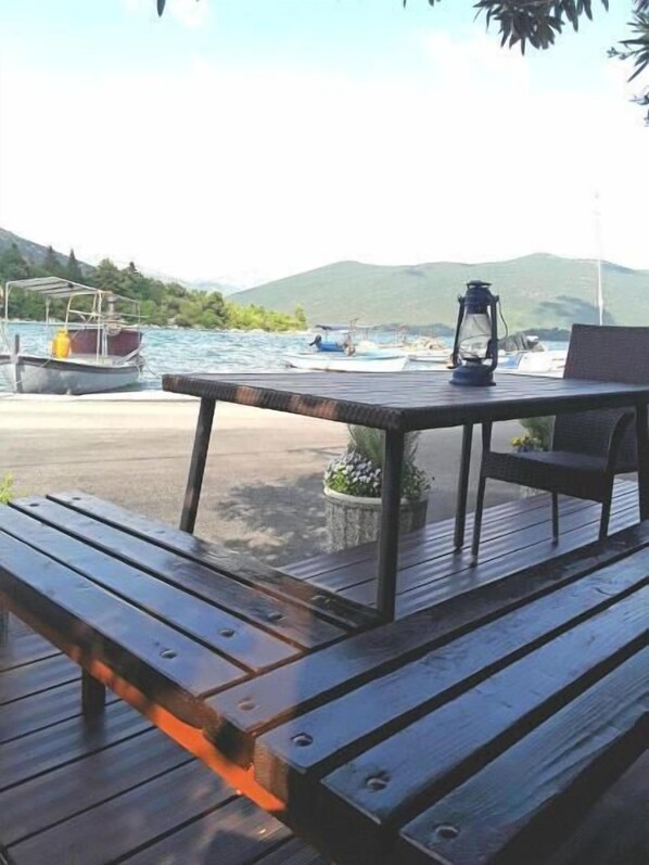 A wooden deck with a table and chairs, overlooking a body of water with boats and hills in the background. - Apartments Nina & Kate (Ston)