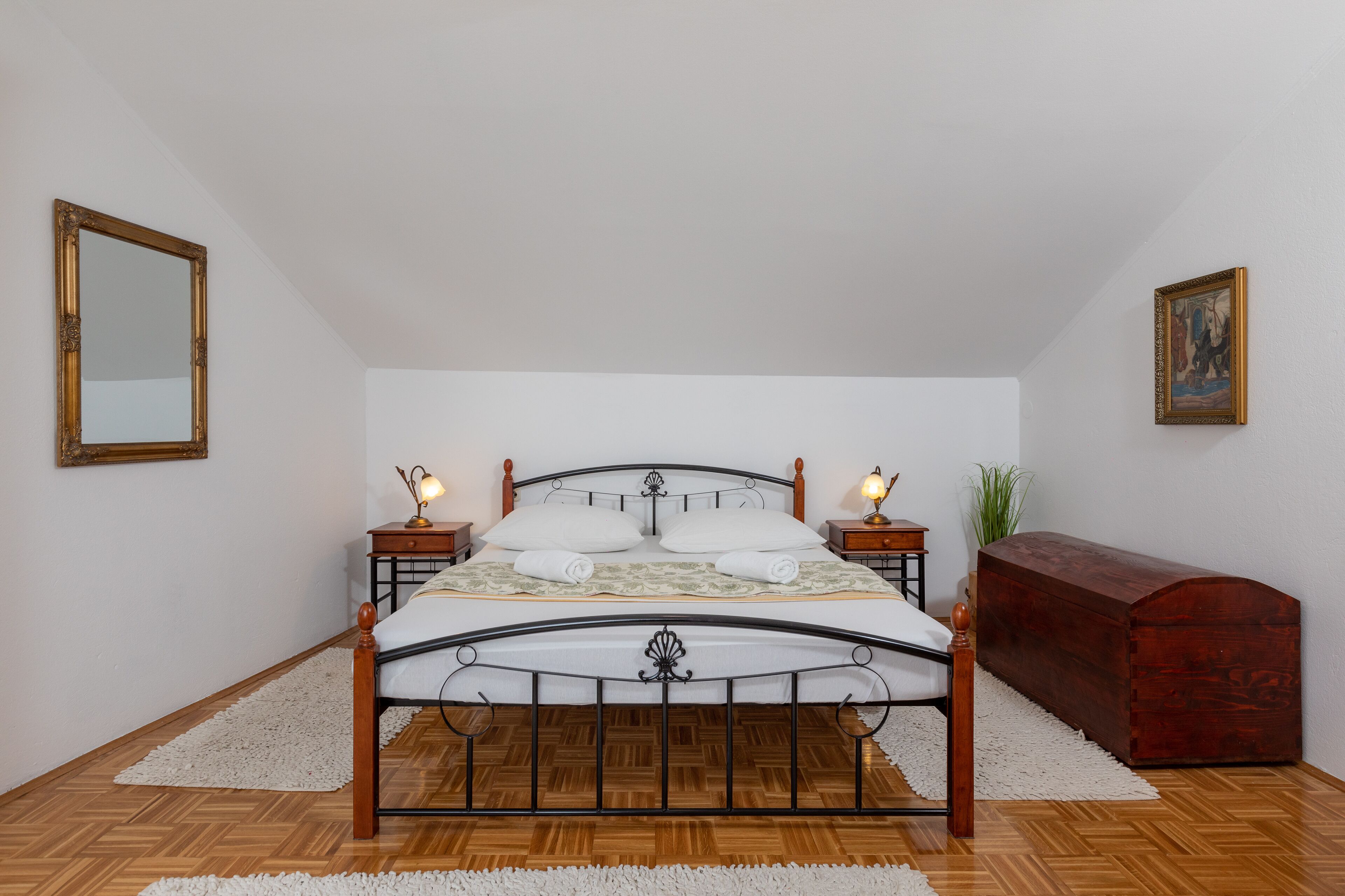 A bedroom with a wooden bed, two bedside tables with lamps, a nightstand, a wooden chest, and a mirror on the wall.