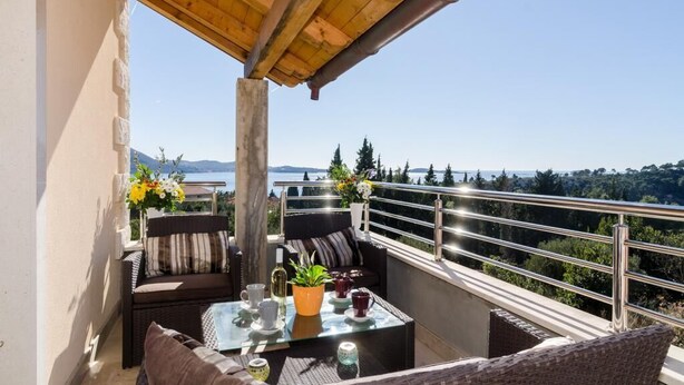 Een balkon met rieten meubels, een glazen tafel en uitzicht op zee en bergen.