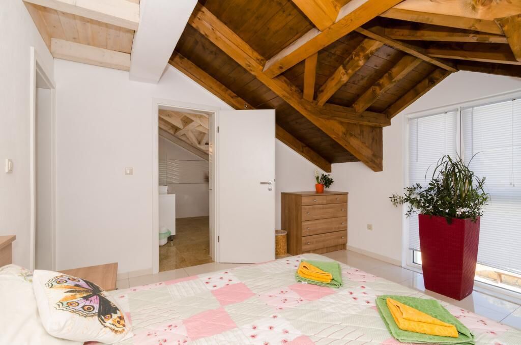 Une chambre avec un plafond en bois, un lit recouvert d’une courtepointe à motifs floraux, une plante rouge et une vue sur une fenêtre.