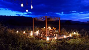 An outdoor wooden gazebo with a table and chairs, surrounded by candles and lanterns, set in a grassy field under a twilight sky. - Asanja Moru (Serengeti)