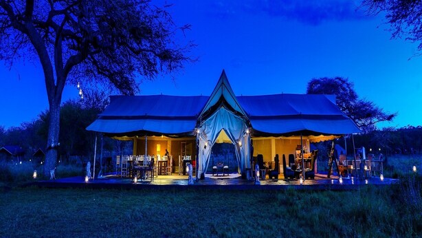 A tented accommodation with a central entrance, surrounded by outdoor seating and tables, set in a natural landscape. - Asanja Moru (Serengeti)