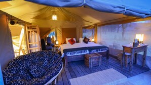 A tented accommodation with a bed, a seating area, and a small table with a lamp. - Asanja Moru (Serengeti)