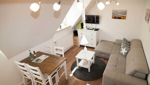 Een gezellige leefruimte met een houten eettafel, witte stoelen, een grijze bank en een klein witte bijzettafeltje.