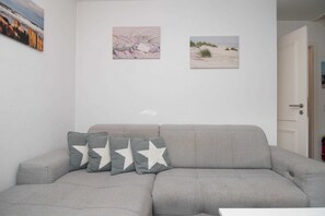 Um sofá cinzento com almofadas estampadas com estrelas, uma porta branca e duas imagens penduradas na parede retratando paisagens.