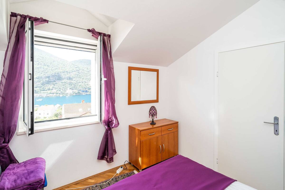 A bedroom with a bed, a wooden wardrobe, a mirror, and a window with purple curtains.