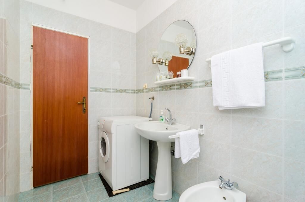 Een badkamer met een houten deur, een wasmachine, een wastafel, een spiegel en een handdoekhouder.
