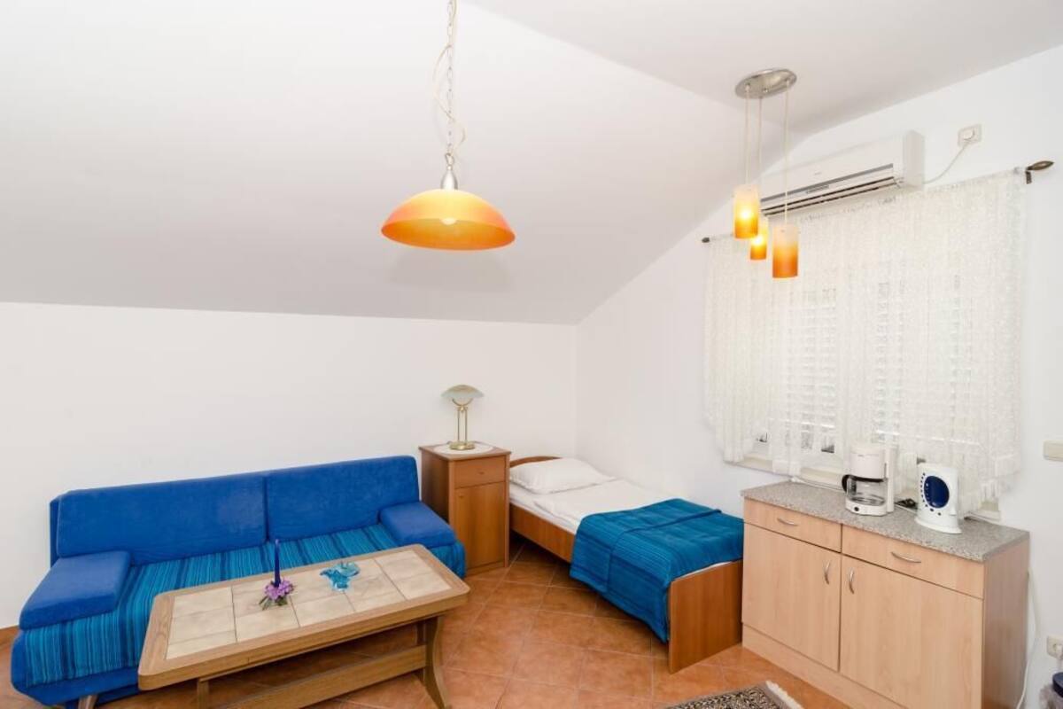 A small, well-lit room with a blue sofa, a wooden table, a bed with a blue bedspread, a small cabinet, and a window with curtains.