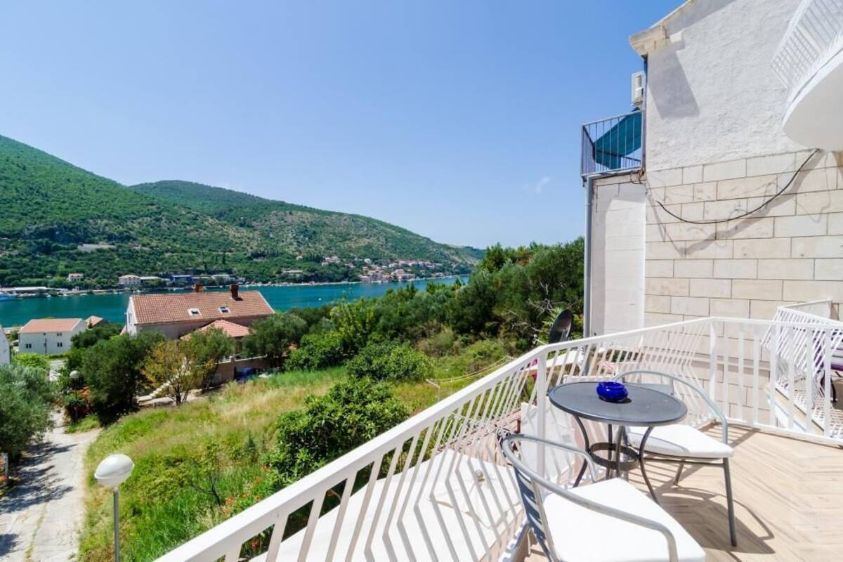 A balcony with a table and chairs overlooking a coastal area with houses and a mountain.