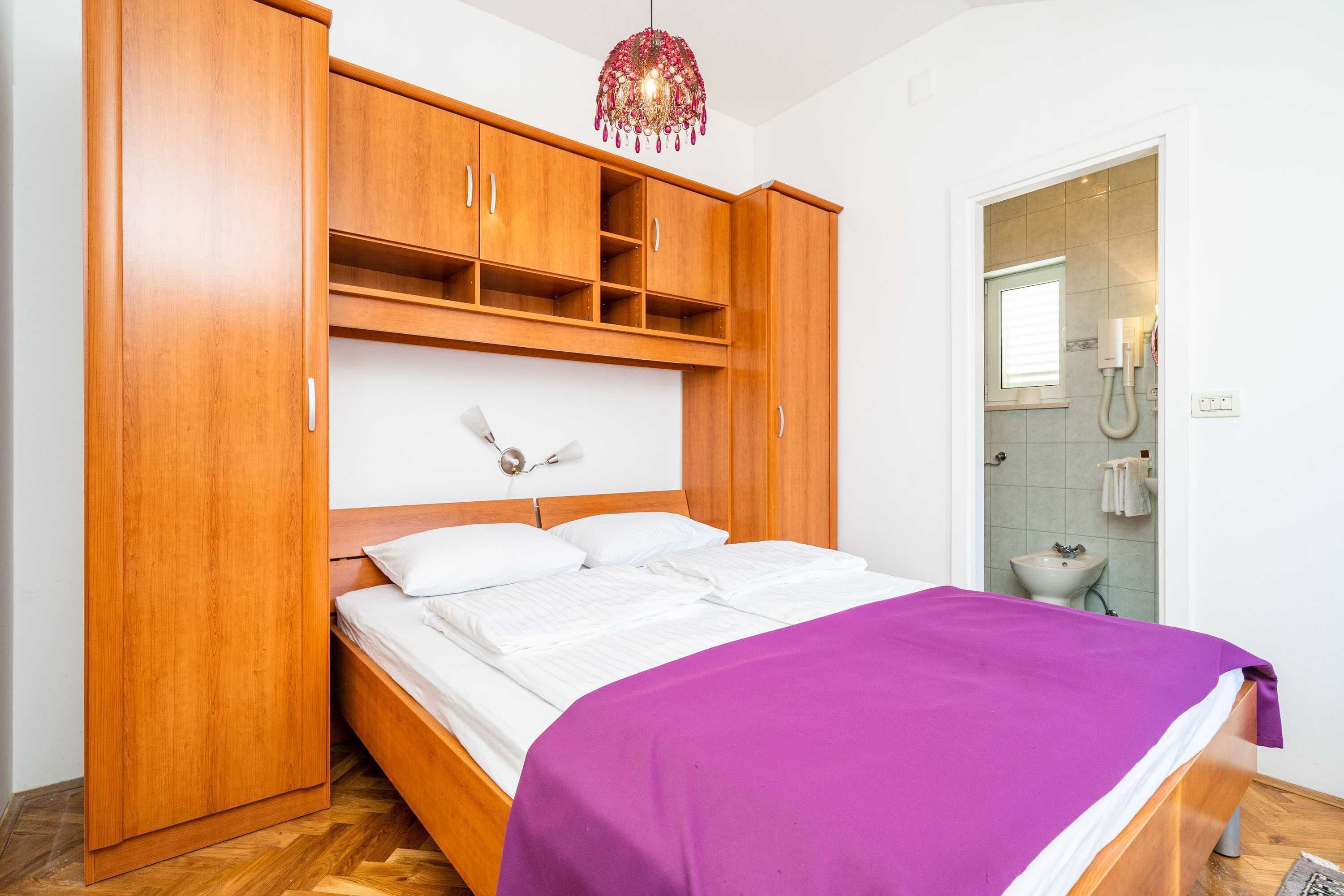 A bedroom with a bed, wooden wardrobe, and a bathroom visible through an open door.