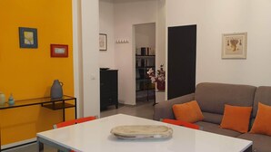 Ein Wohnzimmer mit grauem Sofa, orangefarbenen Kissen, einem weißen Tisch und einem Glastisch. An den Wänden hängen Bilder in Rahmen, und im Hintergrund steht ein schwarzes Sideboard.
