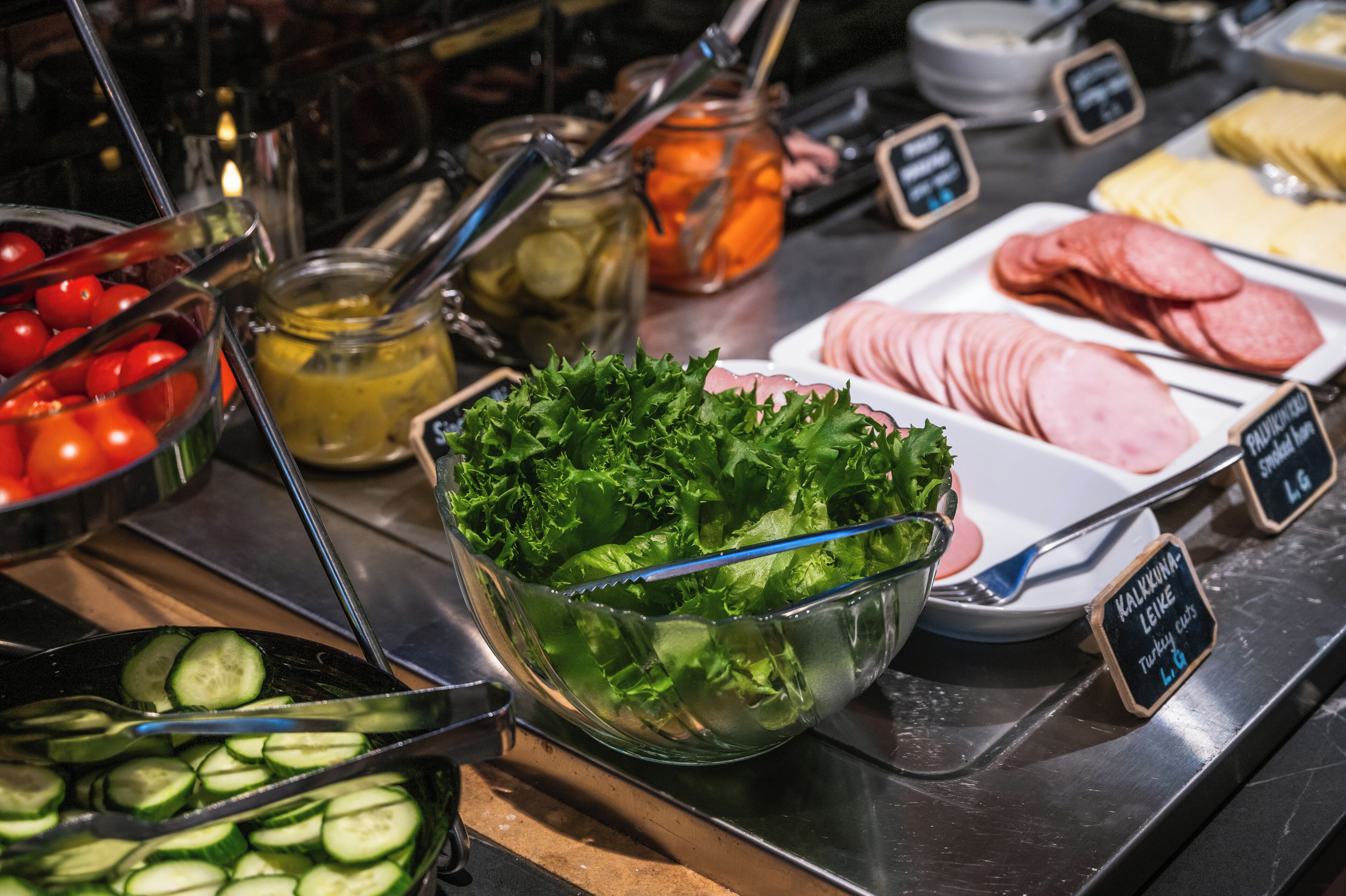 A buffet with various food items including sliced cucumbers, tomatoes, pickles, and different types of meat.