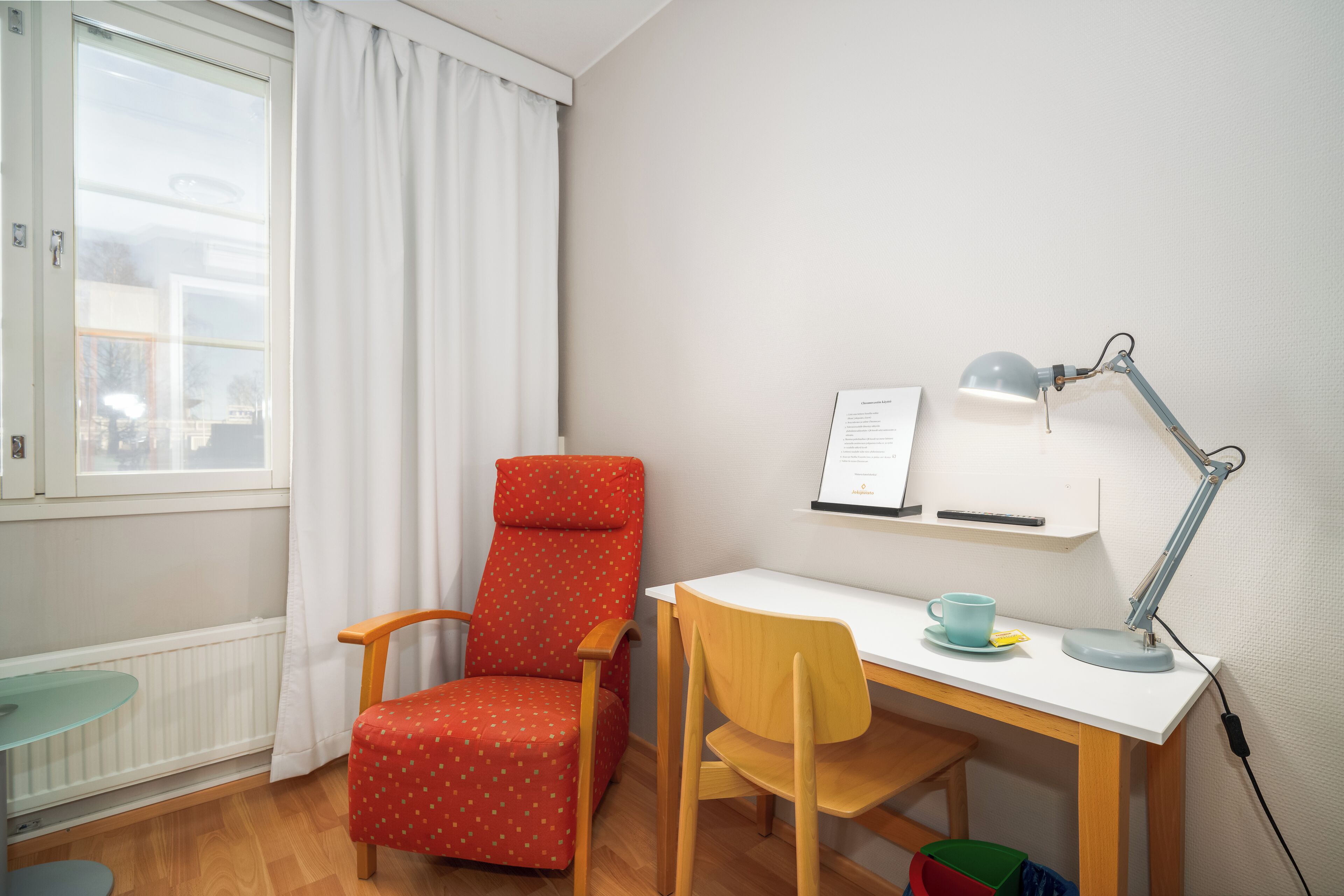 Een kamer met een wit bureau, een rode stoel, een blauw kopje, een witte lamp en een raam met witte gordijnen.