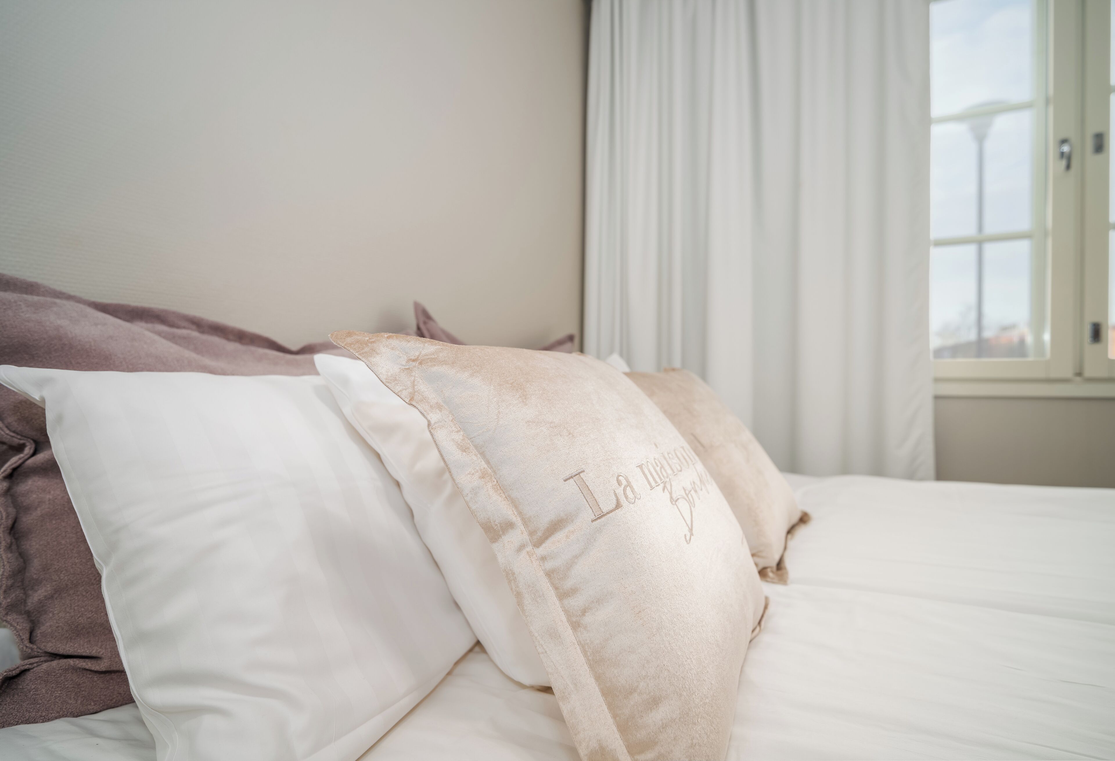 Una cama con almohadas, una ventana con cortinas y vista a un edificio.