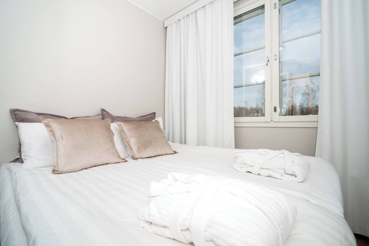 A neatly made bed with pillows, a window with curtains, and a rolled-up towel on the bed.