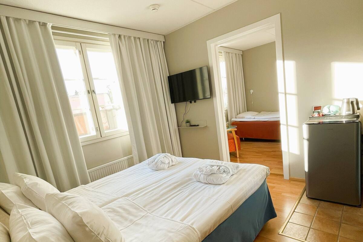 A hotel room with a bed, a TV mounted on the wall, a desk with a computer, and a window with curtains.