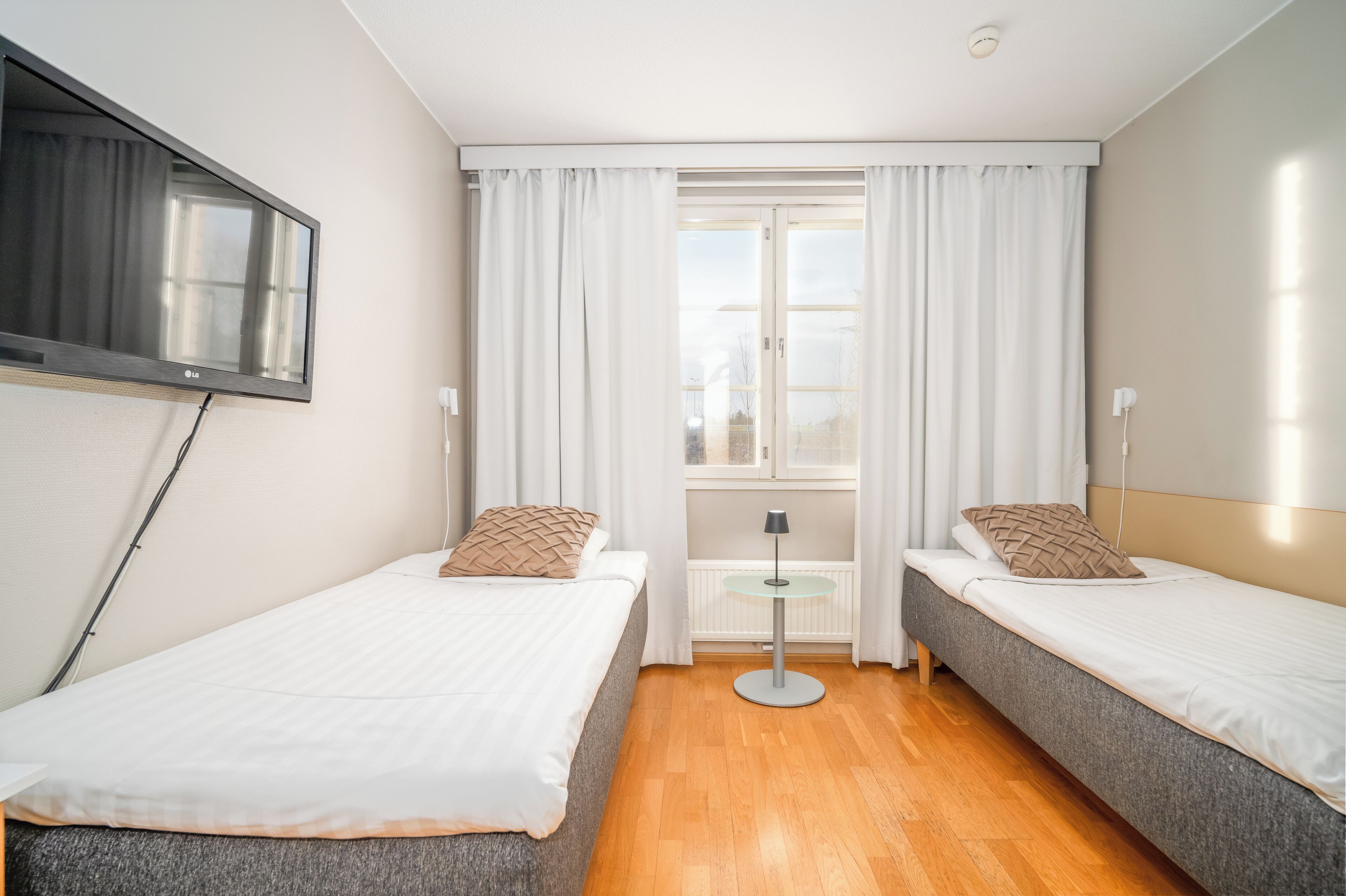 A hotel room with two beds, a TV mounted on the wall, a window with curtains, and a small table with a lamp.