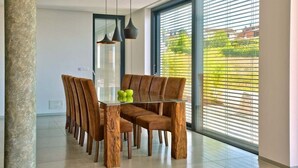A dining area with a glass table and wooden chairs, a stone pillar, and a view through large windows with blinds.