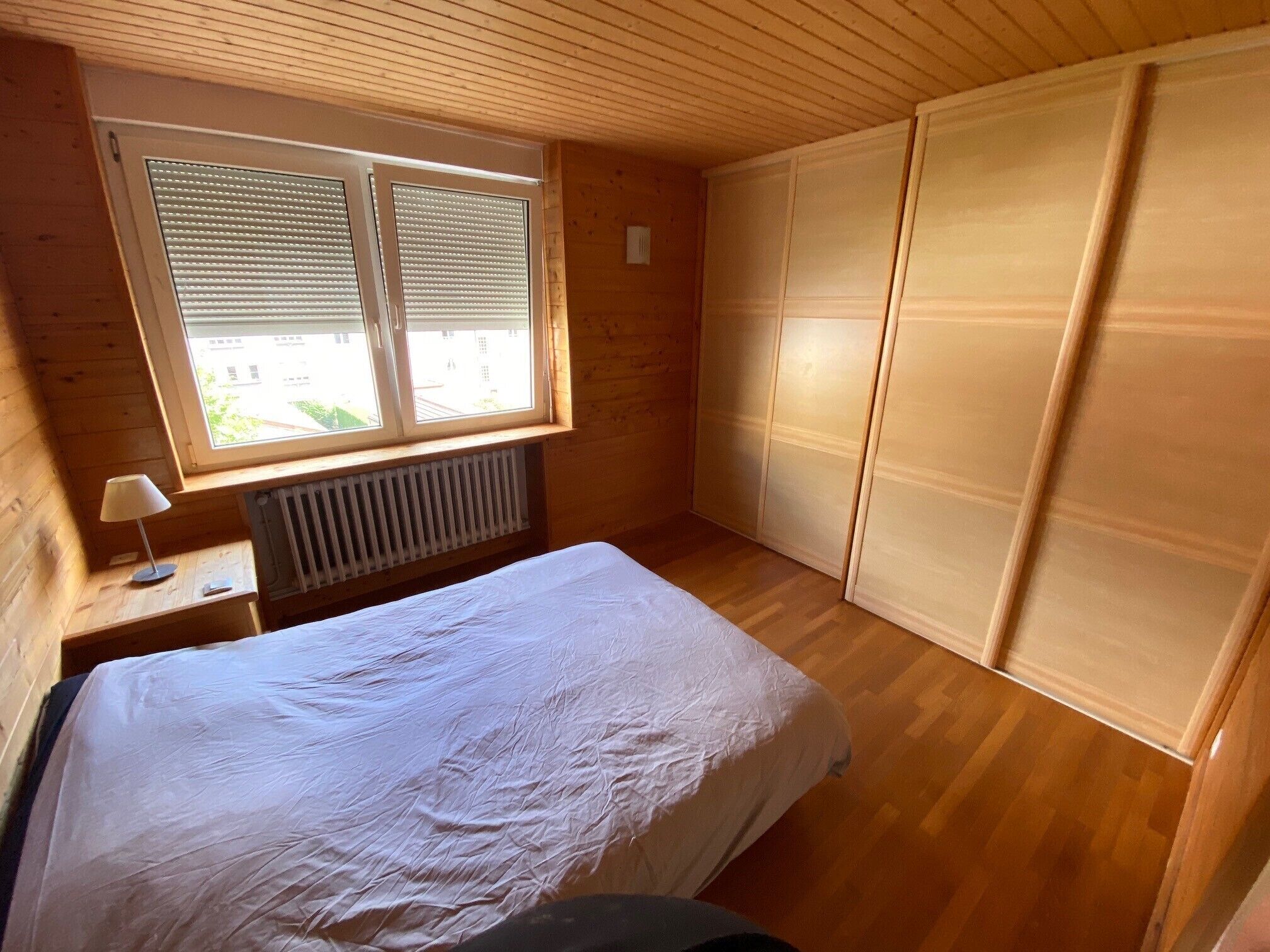 Een kamer met een bed, een raam met zonwering, een radiator en houten wanden.