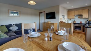Una sala moderna con una mesa de comedor preparada para seis personas, un sofá, un televisor montado en la pared y una cocina con armarios de madera y electrodomésticos.
