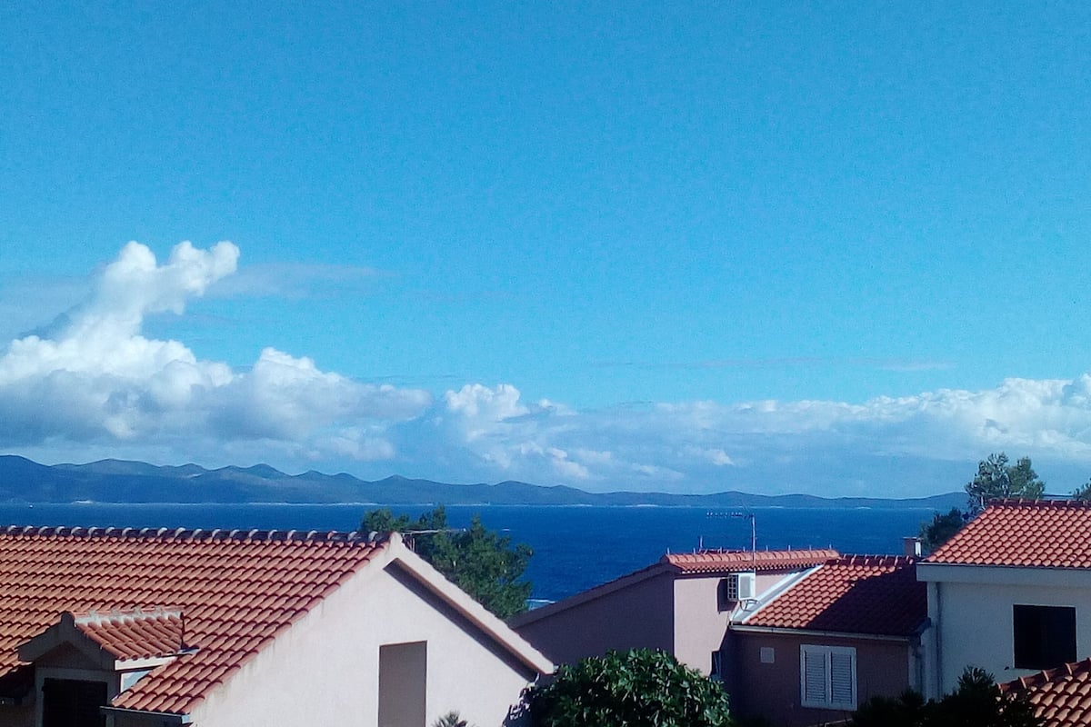 A coastal residential area with red-tiled roofs and a clear view of the sea and distant mountains.
