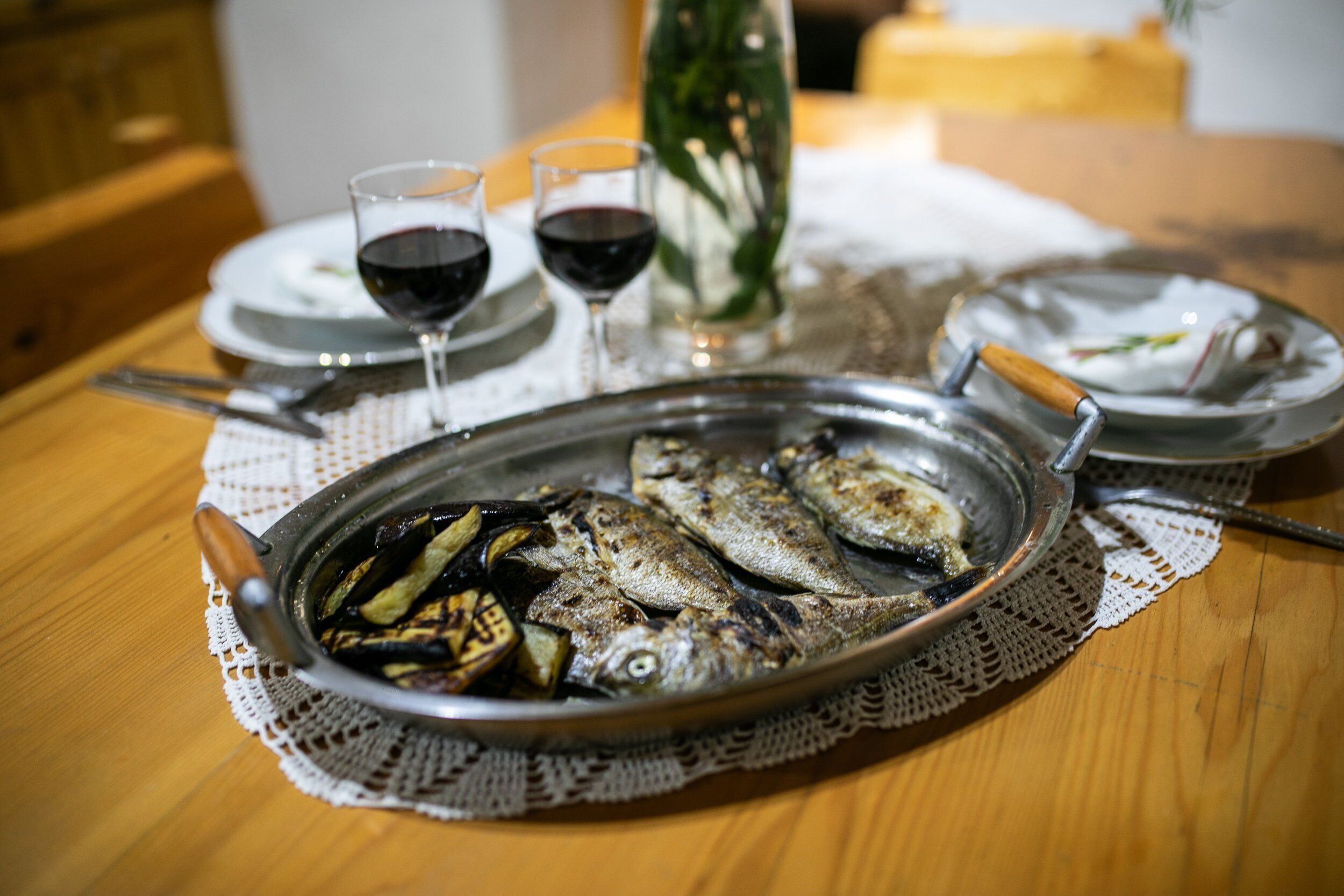 Mesa de jantar com bandeja de metal contendo peixe grelhado, dois copos de vinho e uma garrafa de vinho.