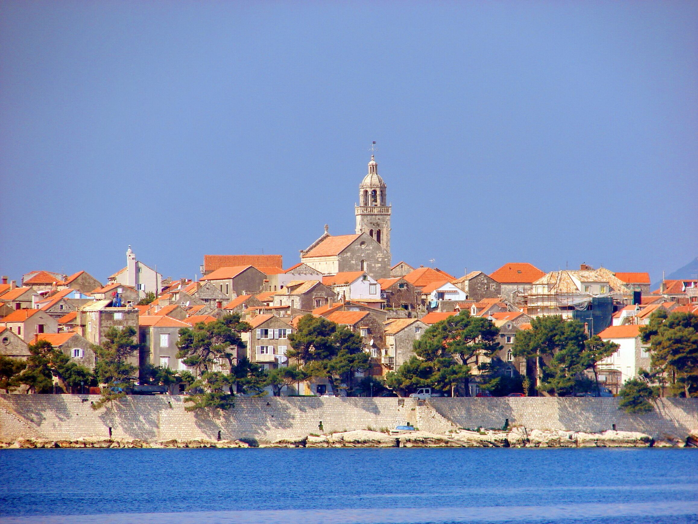 Un pueblo costero con edificios históricos, una imponente torre de iglesia y un muro de piedra a lo largo del paseo marítimo.