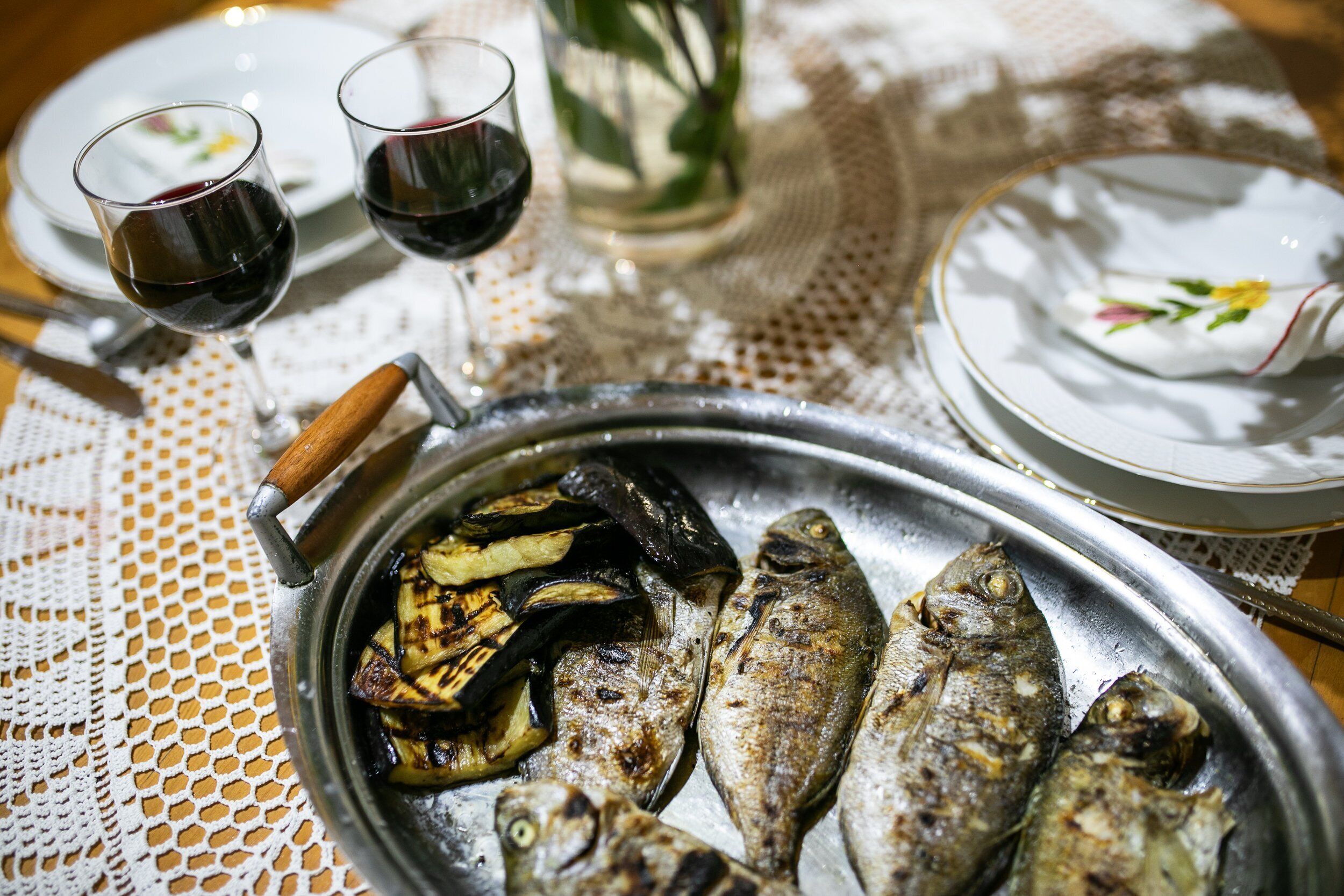 A table with a metal tray containing grilled fish and vegetables, two glasses of red wine, and a white plate with a floral design.