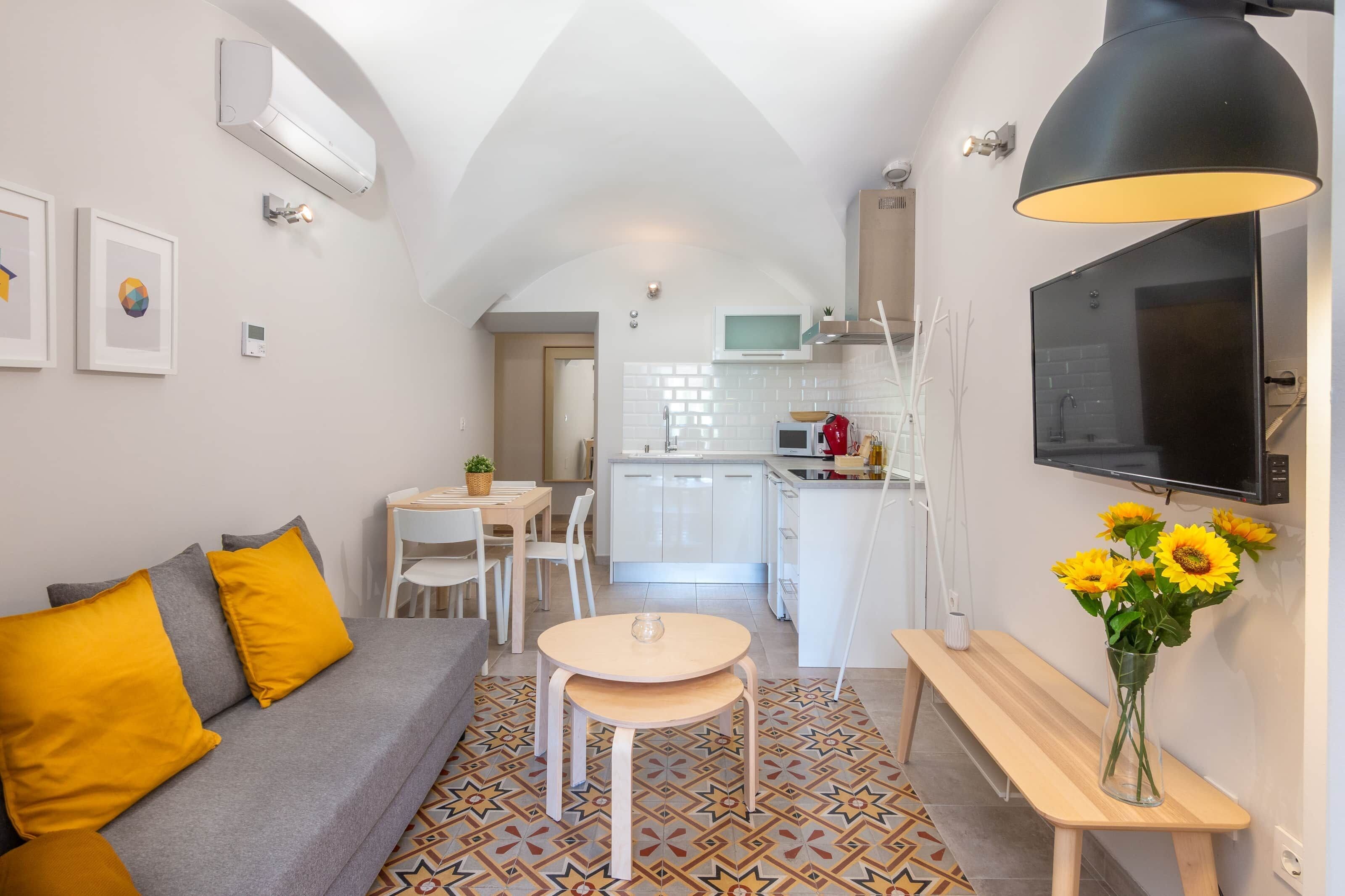 Un espacio habitable compacto con sofá, una pequeña mesa de comedor, un banco, televisor y área de cocina.