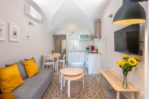 A compact living space with a sofa, a small dining table, a bench, a TV, and a kitchen area.