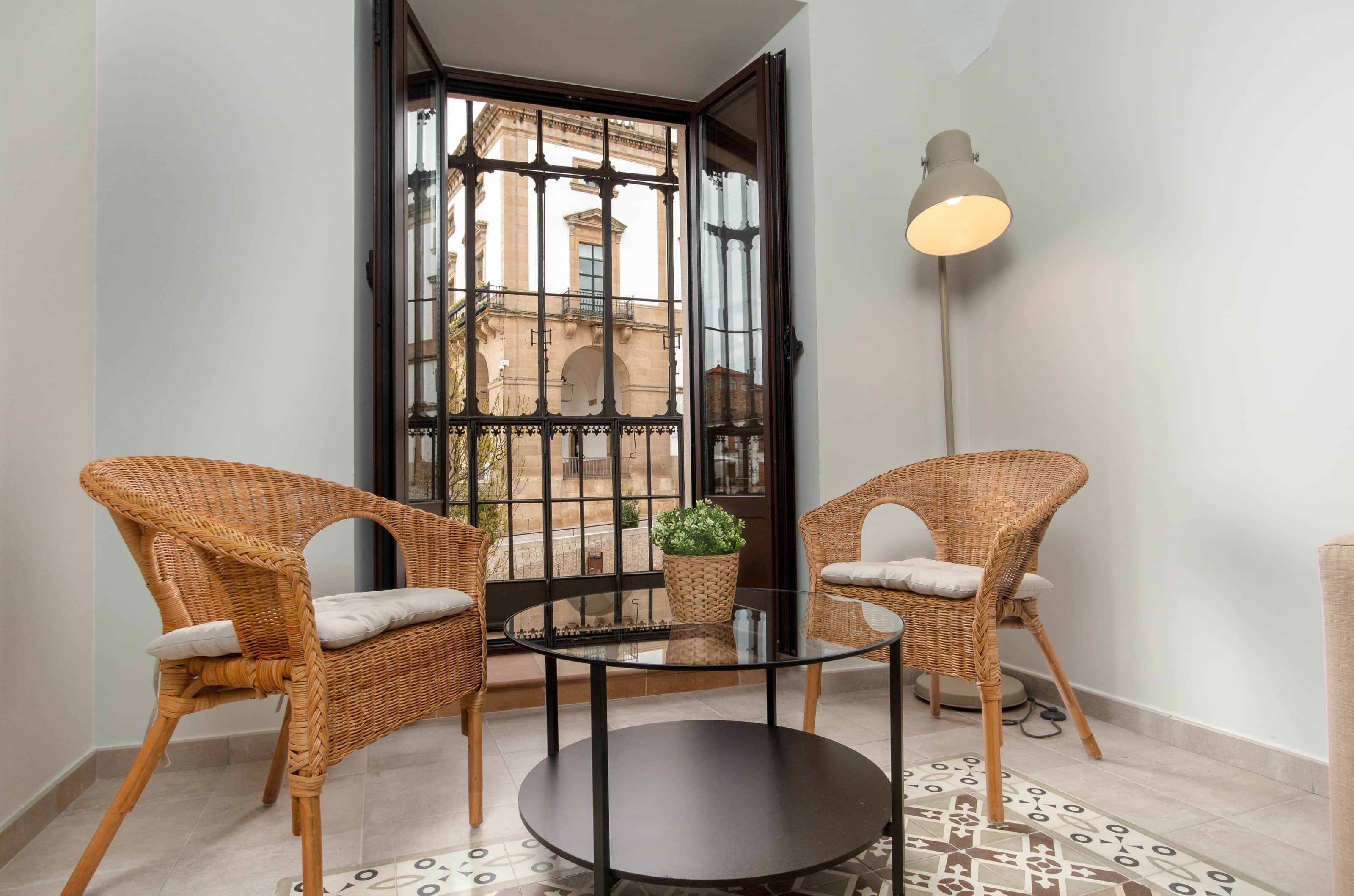 A room with two wicker chairs, a glass table, a floor lamp, and a view through a large window.