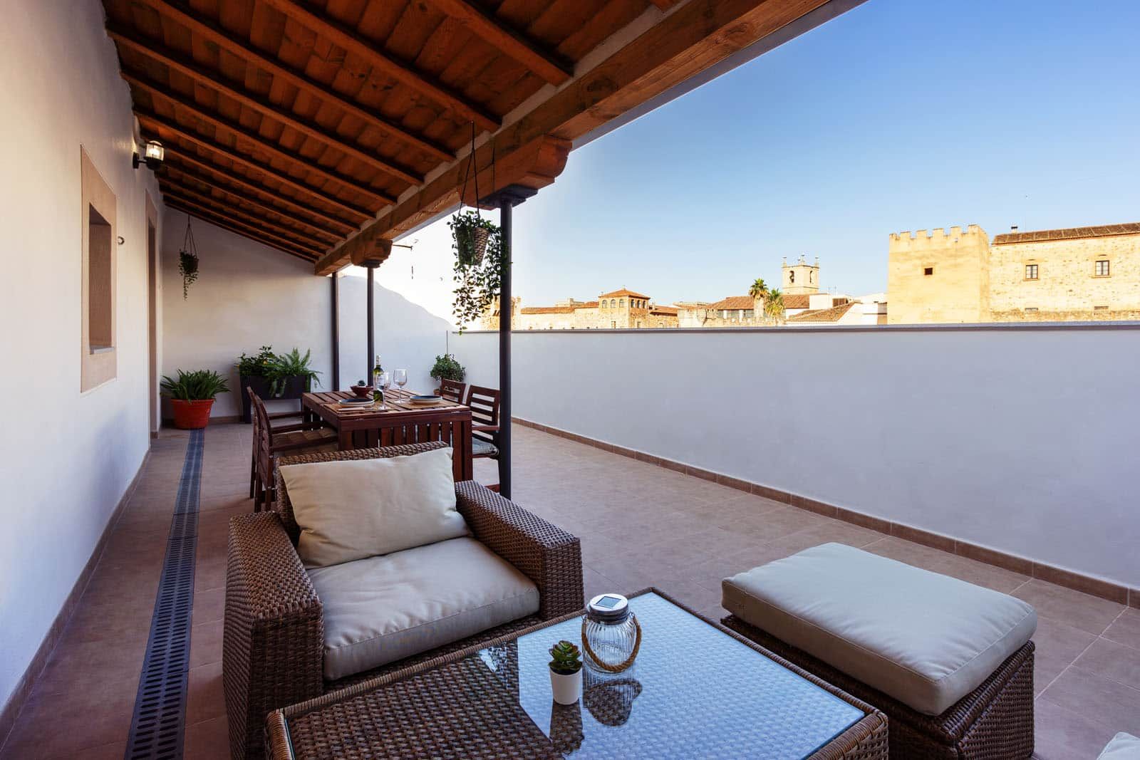 Un espacio de estar techado al aire libre con mobiliario de mimbre y una mesa, con vista a un paisaje urbano.