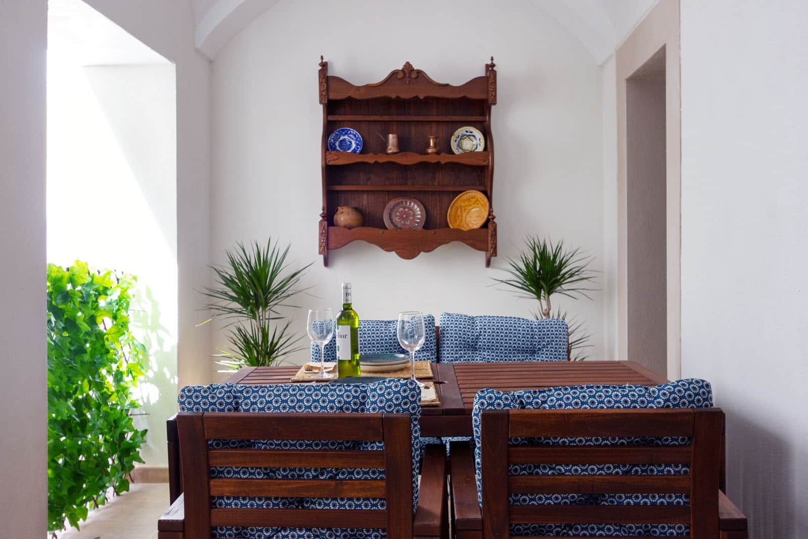Een eetruimte met een houten tafel en stoelen, een wandplank met decoratieve borden, en potplanten.